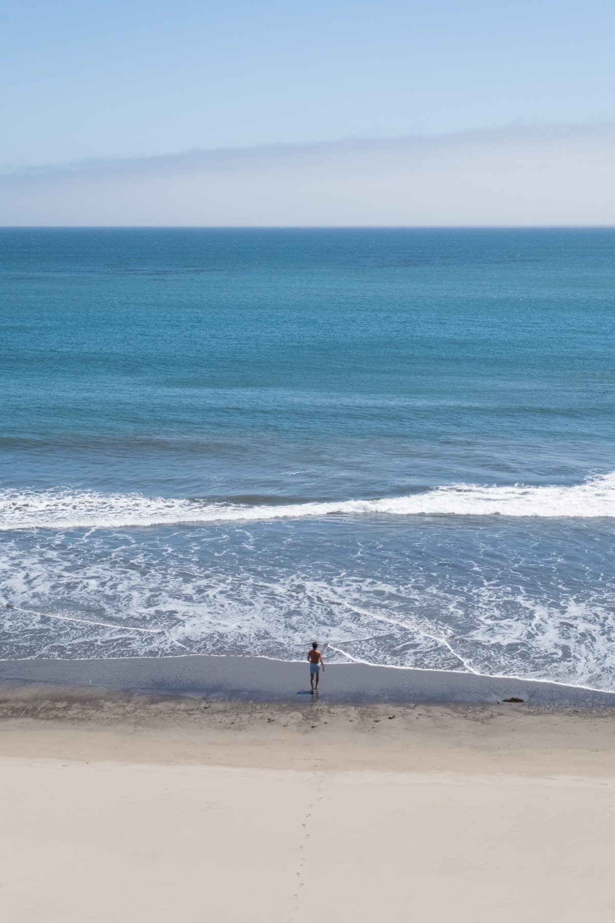 A lone swimmer at the edge of the surf, seen from the bluff above a wide blue ocean